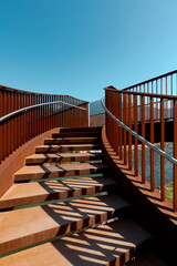Detail of a stairway of a modern rusty bridge, with a clear blue sky behind it. Strong contrast between lights and shadows.