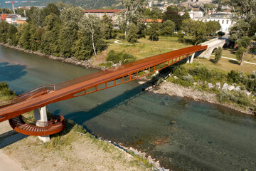 Naklejka na ściany i meble Modern rusted pedestrian and bicycle bridge with flowing Ticino river below. Beautiful panorama of the Alps of Italian Switzerland, taken from the sky
