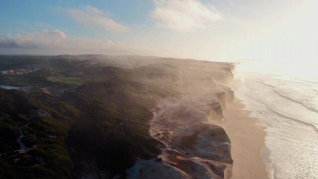 Cinematic drone panning shot over epic rugged coastline of Southern Portuguese coast in Peniche. Inspiring and incredible misty landscape of steep cliffs hanging over empty sand beach with ocean waves