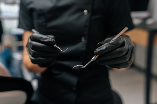 Close-up Cropped Shot Of Unrecognizable Female Practitioner Dentist Wearing Rubber Gloves And Black Uniform Holding Tooth Mirror And Dental Pick, Performing Dental Exam On Patient In Dental Clinic