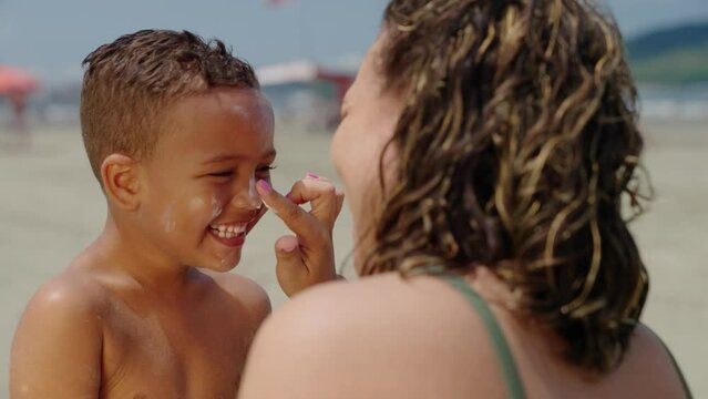mother applying sunblock lotion on sons skin for sun protection little boy getting ready to swim on the beach with mom using sunscreen caring for childs health on sunny day 
