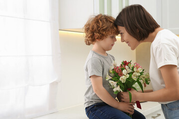 Happy woman with her cute son and bouquet of beautiful flowers in kitchen, space for text. Mother's day celebration