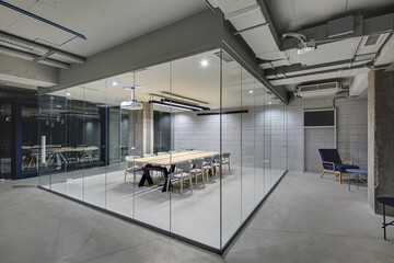 Luminous conference zone in the office in a loft style with brick walls and concrete columns. Zone has a large wooden table with gray chairs and glass walls. Above the table there is a projector.