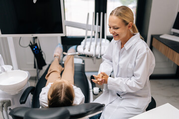 Fototapeta premium Portrait of friendly pediatric dentist bonding with unrecognizable child girl, talking and explaining dental procedure. Back view of little kid learning about healthy teeth, maintaining oral hygiene.