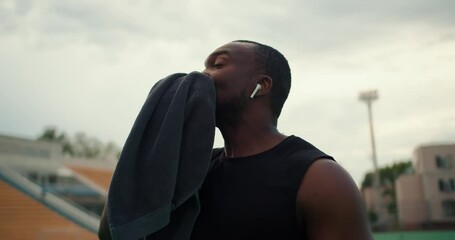 An athlete with black skin in a black T-shirt wipes his face with a towel and drinks water from a special bottle on a city beach against a gray sky
