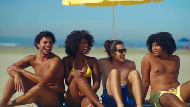 Laughing mixed racial group of friends sitting and relaxing together on the beach in slow motion
