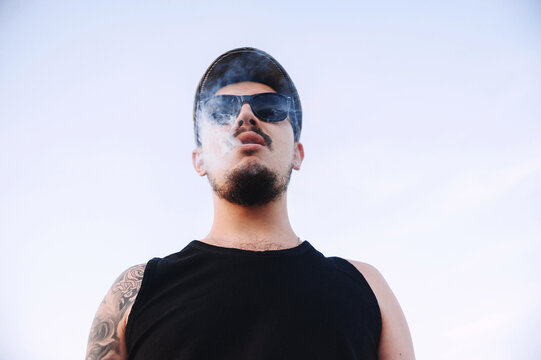 Close-up, portrait of a young guy wearing a cap and sunglasses with a beard and mustache in tattoos, exhaling smoke from a cigarette
