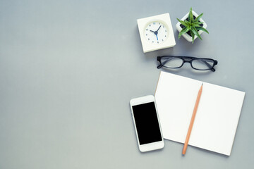 Desk above, table top view of office desk with notebook, pencil, mobile phone, clock, eyeglasses, plant on grey background