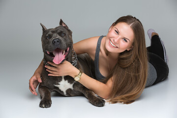 Beautiful sporty young woman lying on floor and hugging adult grey amstafford terrier dog. Studio shot over gray background. Copy space.