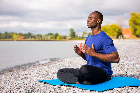 young black man wearing athletic wear sitting on the beach exercising yoga