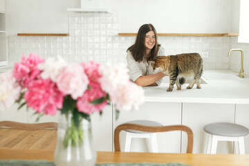 Happy woman caressing cute cat on background of minimal white kitchen with peonies bouquet in new modern home. Housewife relaxing after cleaning up kitchen and playing with pet