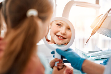 A little girl is a patient at the dentist. A child is holding a mirror and looking at his teeth and smiling.Sunlight.