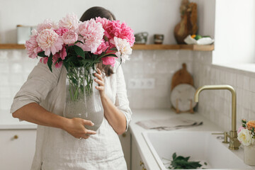 Woman holding beautiful peonies in vase at sink with brass faucet and granite countertop. Stylish female holding pink peony flowers in new modern kitchen, housewife decorating new home © sonyachny