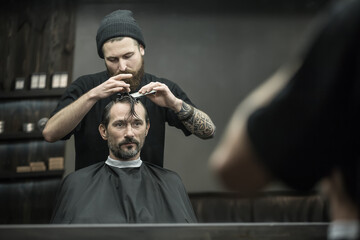 Careful barber with a big beard and a tattoo is combing the hair of the bearded man in the black hair cutting cape in the barbershop. He also fixes client's strands with the hairgrips. Horizontal. - obrazy, fototapety, plakaty