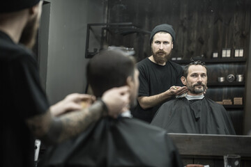 Man and a barber reflected in a mirror in the barbershop. Hairdresser wears a black T-shirt with a cap and has a hair comb. Client wears a black cutting hair cape and has hairgrips on his head. - obrazy, fototapety, plakaty