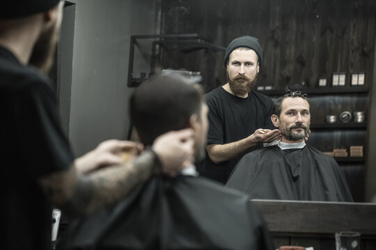 Man and a barber reflected in a mirror in the barbershop. Hairdresser wears a black T-shirt with a cap and has a hair comb. Client wears a black cutting hair cape and has hairgrips on his head.