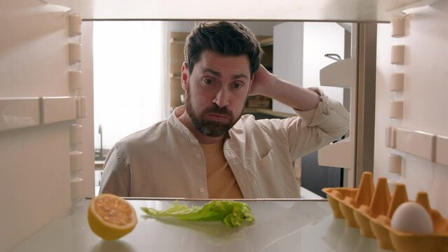 Hungry man guy open empty refrigerator with half lemon lettuce leaf one egg slice of bread leftover orange juice in bottle confused puzzled sad need food delivery point of view POV from inside fridge