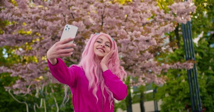 A happy girl in pink makes a selfie on the background of a tree that blooms with pink flowers. Selfie on a walk, blog, happy moments on camera