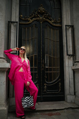 Fashionable woman wearing trendy pink fuchsia color linen suit with oversized shirt, wide leg trousers, sunglasses, holding zebra print tote bag, posing in street of city. Full-length outdoor portrait