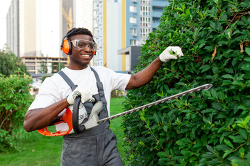 african american male garden worker in uniform trims bushes with electric tool