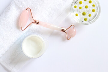 Fototapeta premium Rose quartz facial roller. Facial massage roller, chamomile water, cream and white towel. Flat lay on a light background. Top view, copy space. Spa