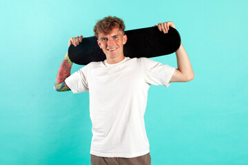 young cheerful tattooed guy in white t-shirt holding skateboard and smiling on blue isolated background © Богдан Маліцький
