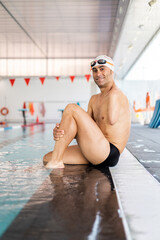 An adult swimmer with an amputated arm sits on the edge of a heated indoor pool with a hat and goggles while looking at the camera. Concept of disabled athletes, swimmers with an amputated arm.