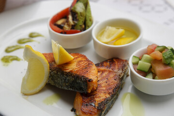 Fried salmon steak served with grilled vegetables, olive oil, lemon slices and pesto sauce on the restaurant table