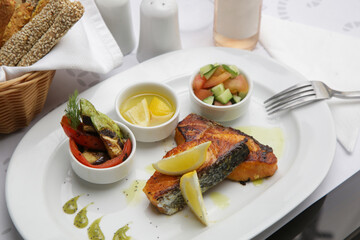 Fried salmon steak served with grilled vegetables, olive oil, lemon slices and pesto sauce on the restaurant table