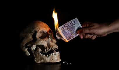 Burning banknote of 500 euros in a man's hand and a human skull on a black background