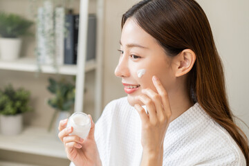 Facial beauty skin care, smile of pretty asian young woman in bathrobe looking at mirror, hand applying moisturizer lotion on her face, holding jar of skin cream before makeup cosmetic routine at home - obrazy, fototapety, plakaty