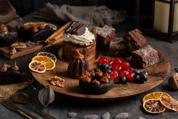 cakes puddings and chocolate muffins with blueberries and cream cherries and slices of dried citrus on highly decorated wooden plate.