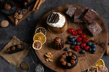 cakes puddings and chocolate muffins with blueberries and cream cherries and slices of dried citrus on highly decorated wooden plate.