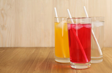 Glasses of different refreshing soda water with ice cubes and straws on wooden table. Space for text