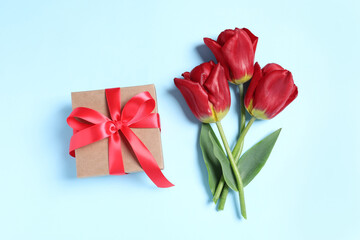 Beautiful gift box with bow and red tulips on light blue background, flat lay