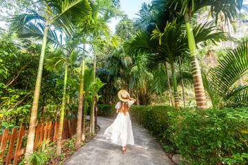Back view woman in white dress straw hat walks along tropical path in Thailand resort. Concept of tropical vacation and nature beauty.
