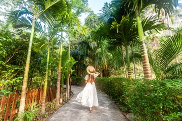 Fotobehang Back view woman in white dress straw hat walks along tropical path in Thailand resort. Concept of tropical vacation and nature beauty. © TravelMedia