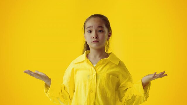 Portrait of a asian kid girl gesturing with raised hands, shrugs his shoulders, being buffled and confused, posing over yellow color background wall in studio