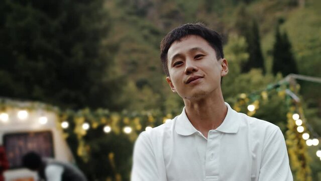 Portrait of a young asian guy at outdoor party relaxing and enjoying music. Smiling and posing on a camera. Trees on a background