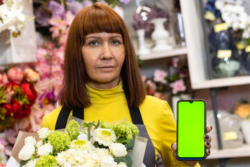 Floristry. Flowers. A florist girl with a bouquet of flowers, and with a phone with a green screen, chromakey. Shop, against the background of a showcase with garden decor.