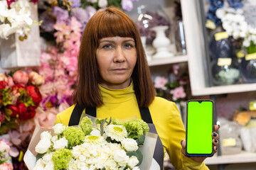 Floristry. Flowers. A florist girl with a bouquet of flowers, and with a phone with a green screen, chromakey. Shop, against the background of a showcase with garden decor.