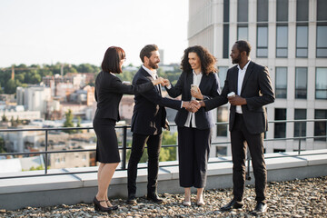 Multicultural team of specialists shaking hands and expressing satisfaction from holding efficient meeting on fresh air. Four diverse colleagues reaching mutual agreement for continued partnership.