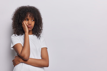 Fototapeta na wymiar Photo of dissatisfied curly haired woman keeps hand on cheek concentrated aside dressed in casual t shirt isolated over white background copy space for your promotional content or advertisement