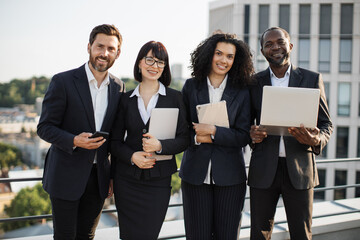 Portrait of happy entrepreneurs of caucasian and african american nationality standing next to each other and smiling at camera. Positive attractive business partners holding modern gadgets in hands.