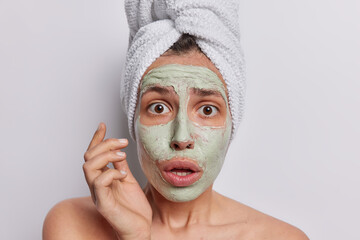 European woman in state of stunned amazement her eyes wide open and mouth agape engaging in skin care procedures meticulously applies facial beauty mask wears bath towel on head isolated on white wall