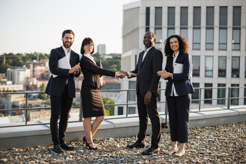 Side view of multiethnic entrepreneurs celebrating contract agreement of becoming partners. African american man and caucasian short haired lady shaking hands while their colleagues clapping on roof.