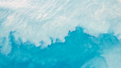 Top view of light blue water with white sand. Waves raise sand and a mix with turquoise water is...