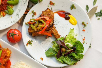Cooked lamb served on a white plate with vegetables, salad, tomatoes, and sauces