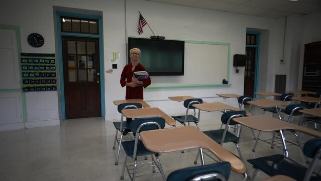 Happy smiling teacher looking longingly at her empty classroom turns and walks out of the room turns off lights. Room has US American flag and smart board with empty desks.