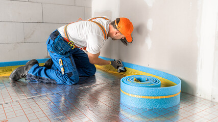 A roll of blue dilatation tape laid on the underfloor insulation. Soft focused Worker on the background during sticking tape to the wall.